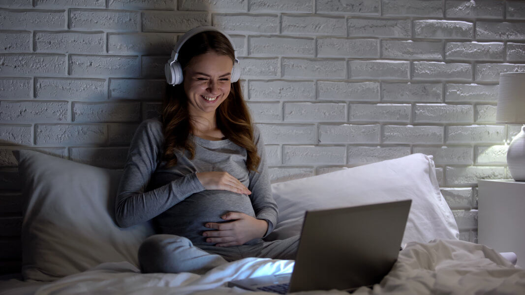 Happy pregnant woman laughing watching comedy on laptop, relaxing in evening