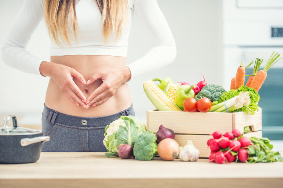 Woman with sports figure on her belly shows heart shape in home kitchen with wooden box full of organic vegetable.