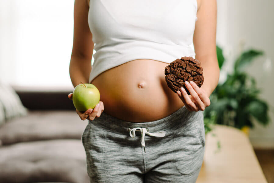 Pregnancy healthy diet and gluttony concept. Woman holding apple and chocolate cookie at home. Pregnancy and sugar.