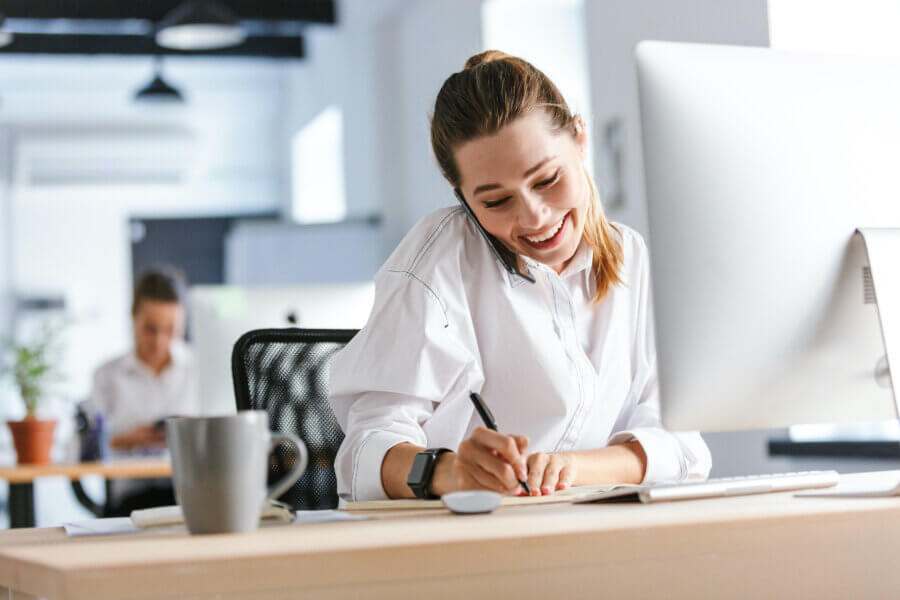 Cheerful young businesswoman sitting at her workplace at the office, talking on mobile phone, taking notes