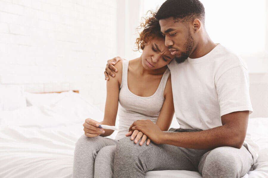 grief Infertility problem. Upset african american couple sitting on bed with negative pregnancy test result, free space